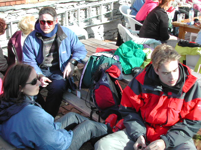 A group of people in winter jackets sit outdoors on benches, some talking and others looking away.