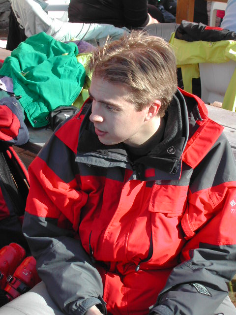 A person in a red and black ski jacket sits outdoors, looking to the side with a neutral expression.