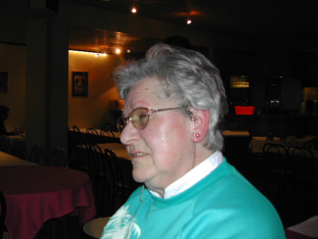An older woman with glasses and a hearing aid sits in a dimly lit restaurant, looking to the side.