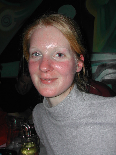 A person with light skin and red cheeks is smiling at the camera, wearing a gray turtleneck sweater indoors.