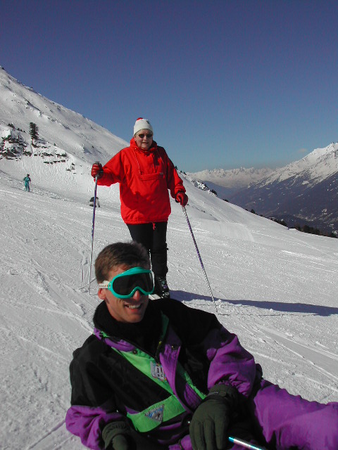 Two people on a snowy mountain; one is skiing, and the other is sitting wearing ski goggles and winter gear.