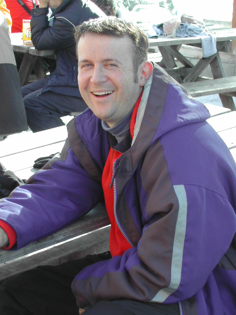 A man in a purple and red jacket sits at a picnic table, smiling at the camera.