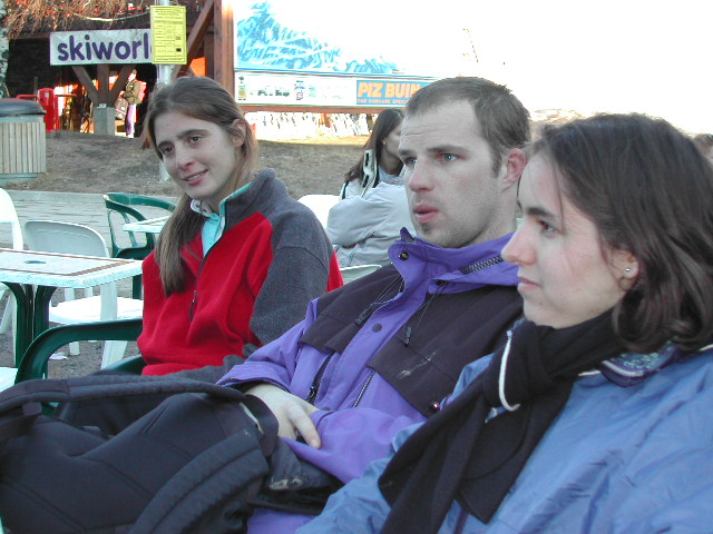 Three people in winter jackets sit outdoors, engaged in conversation, with a ski resort sign visible in the background.