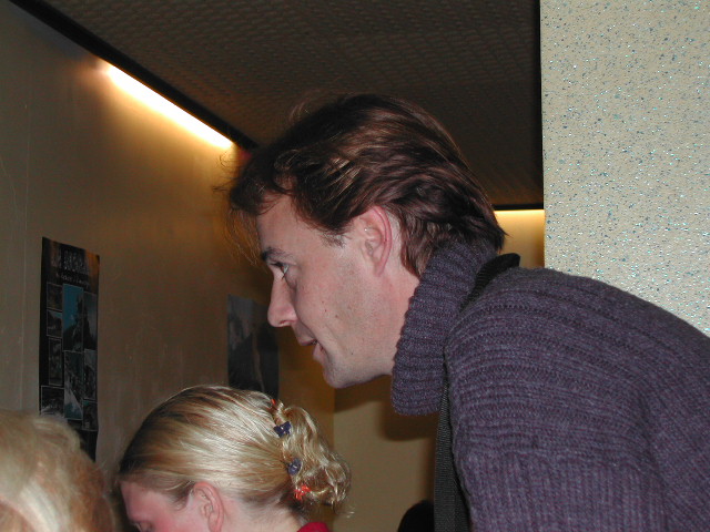 A man in a dark sweater leans forward, looking at something with two people in front of him.