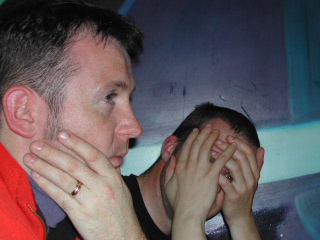 Two men sitting close together; one looks to the side, while the other covers his face with his hands.