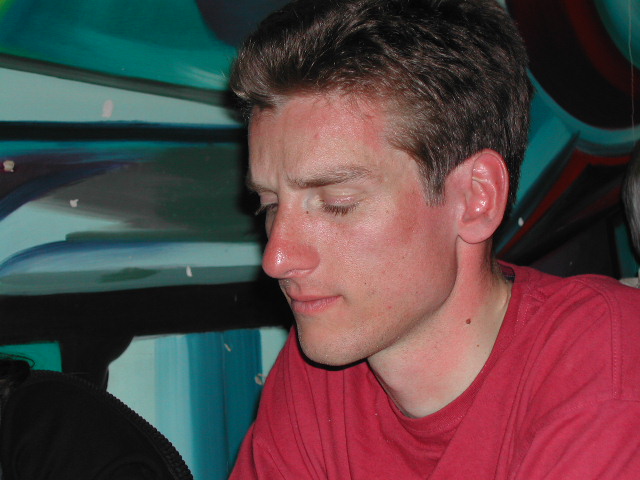 A man in a red shirt sits with his eyes closed, appearing focused or deep in thought.