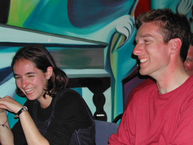 A woman and a man sit together, both laughing and smiling in a colorful indoor setting.
