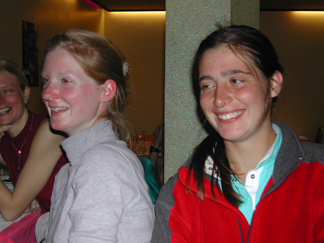 Two women are sitting close together, smiling and laughing, while another person in the background also smiles.