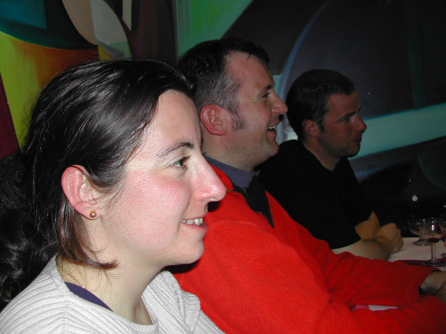 Three people sit together at a table, smiling and engaged in conversation in a dimly lit setting.
