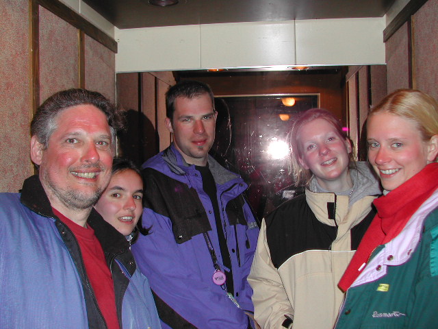 Five people in winter jackets stand close together in a narrow hallway, smiling at the camera.