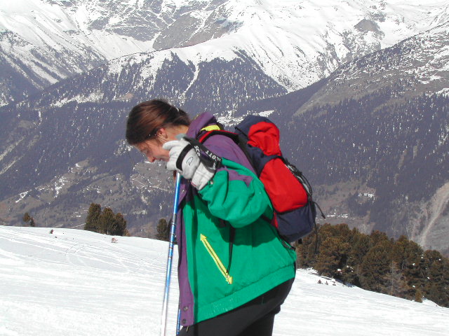 A person in a green jacket and backpack holds ski poles while looking down on a snowy mountain slope.