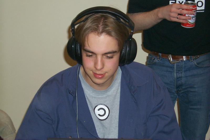 A young person wearing headphones focuses on a computer screen at a LAN party, while another holds a drink nearby.