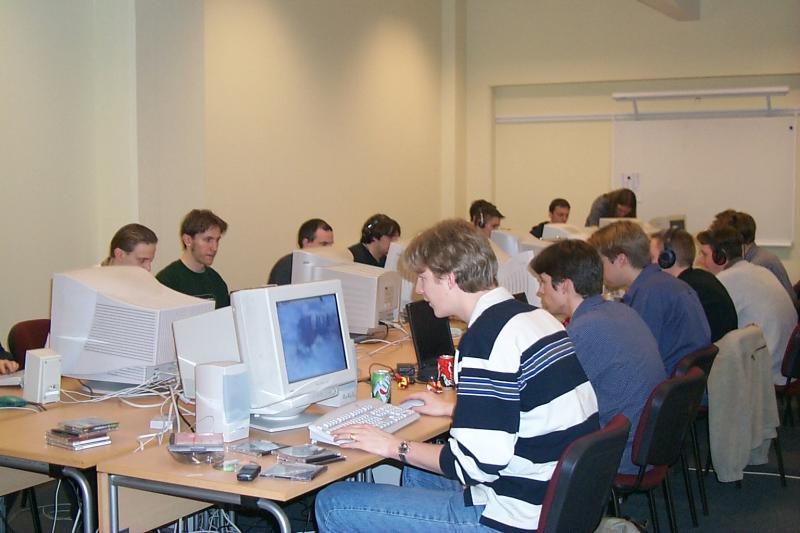 A group of people at a LAN party, sitting at desks with computers, playing games and wearing headphones.