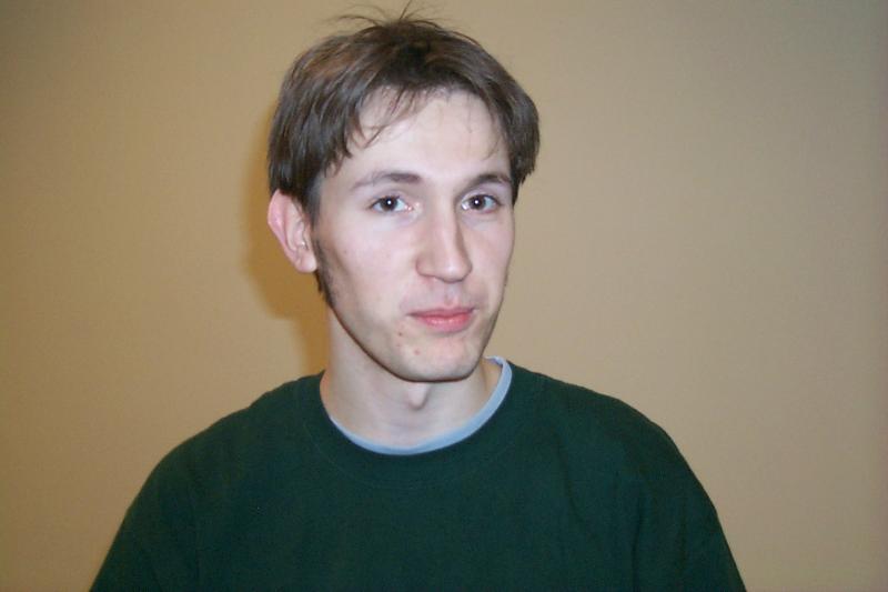 A person with short brown hair wearing a dark green shirt looks at the camera with a neutral expression.