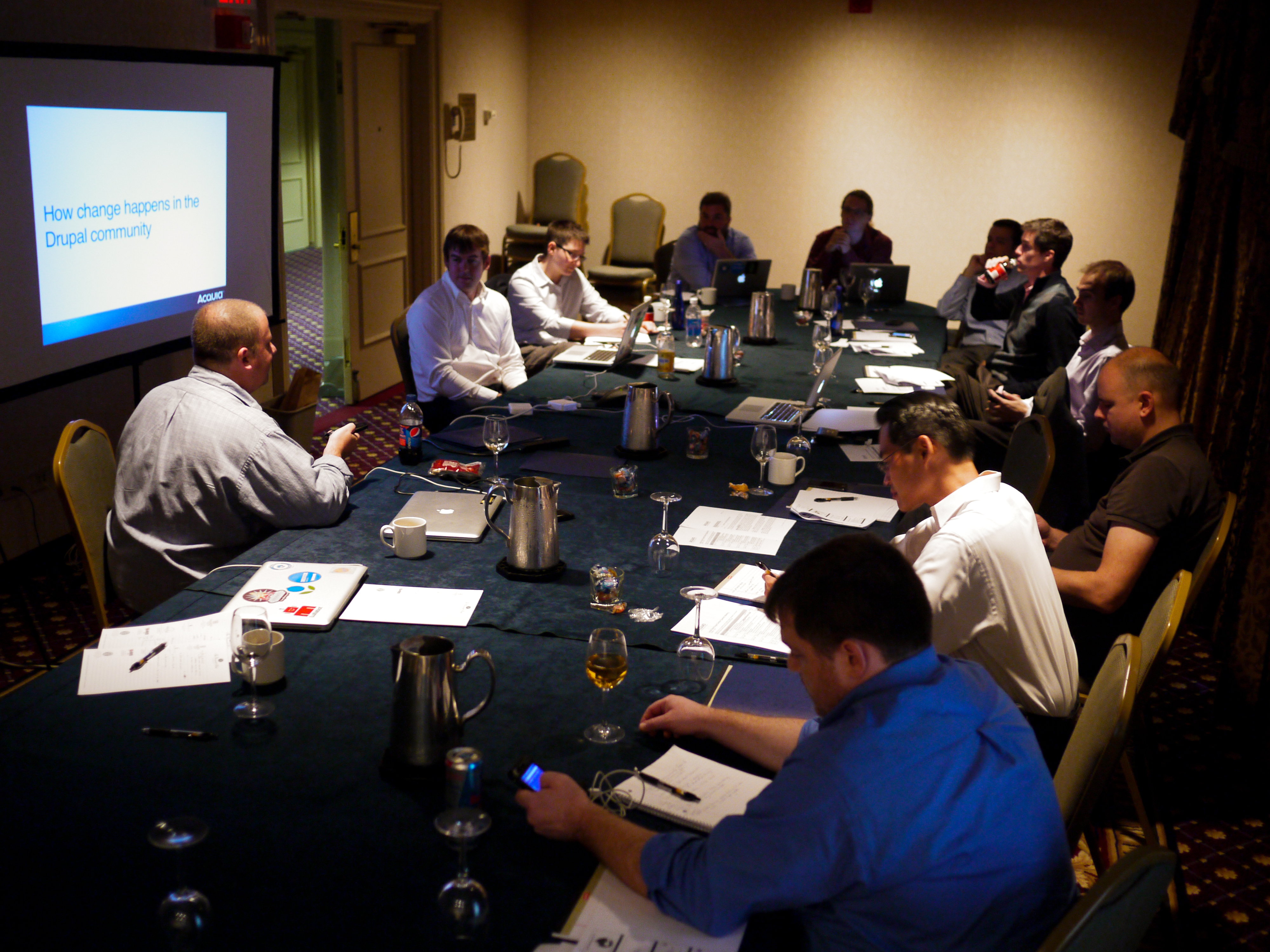 A group of people in a conference room discussing Drupal, with a presentation displayed on a projector screen.