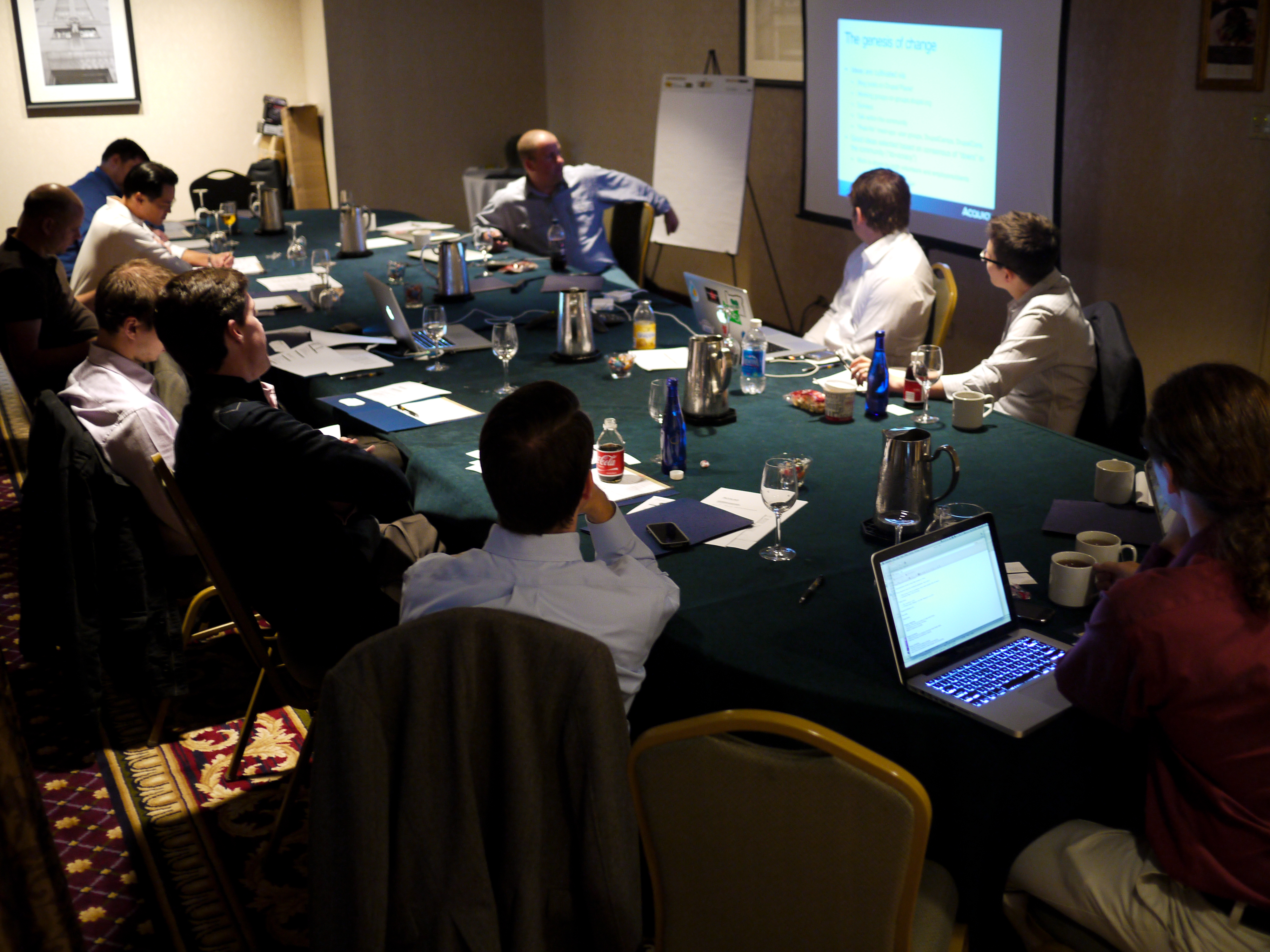 A group of people in a conference room listen to a presenter speaking in front of a projected screen.