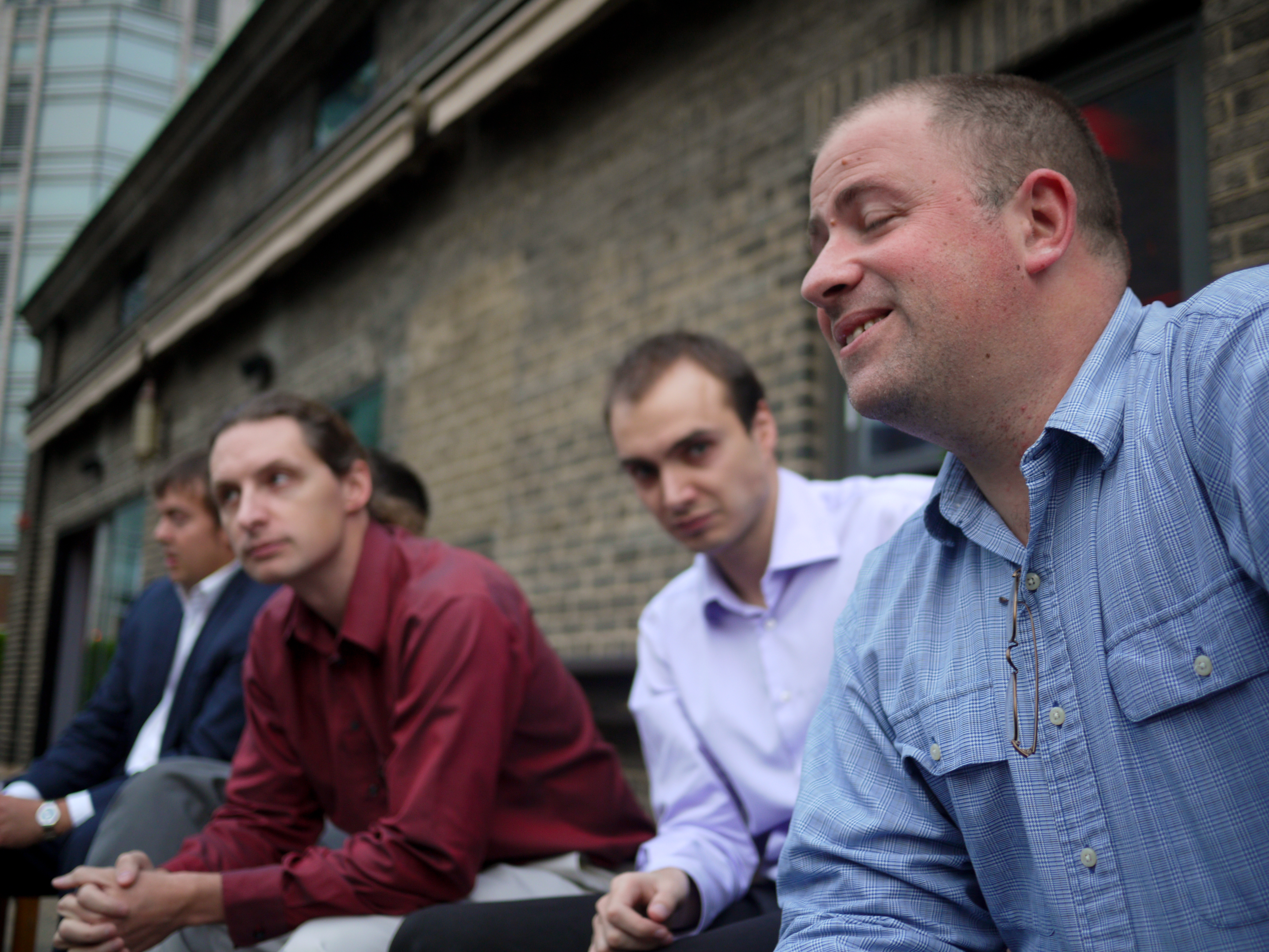 Four men sit outside near a brick building, with one in a blue shirt smiling and speaking.
