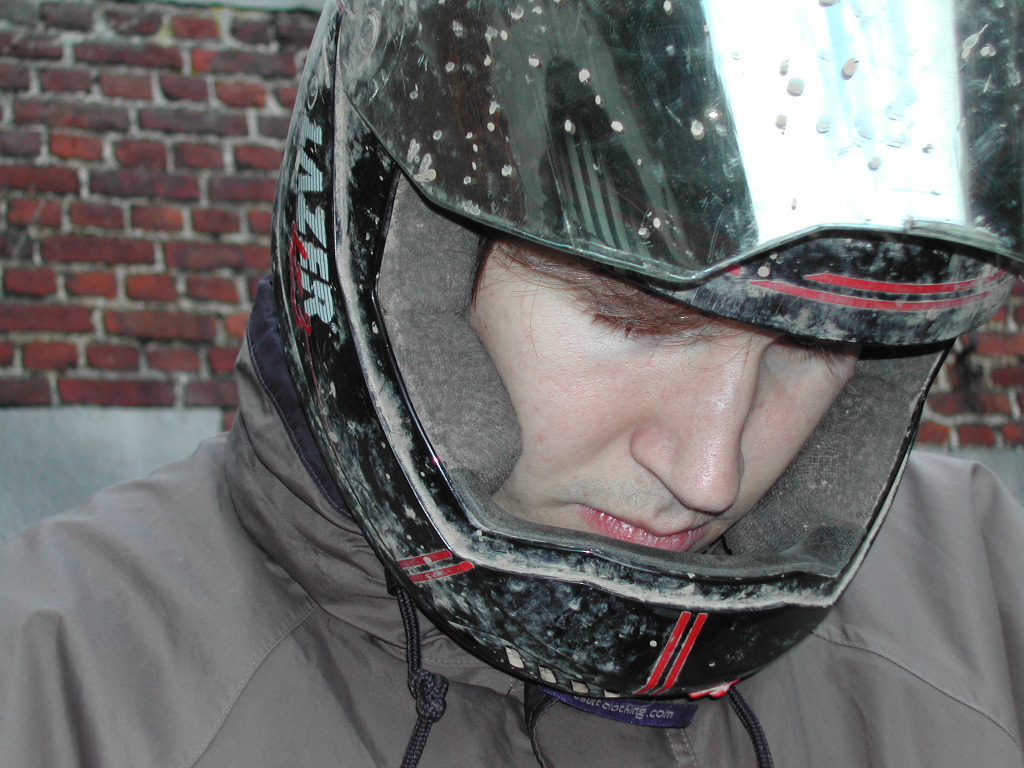 A person wearing a scratched motorcycle helmet looks downward, with a reflective visor partially covering their face.