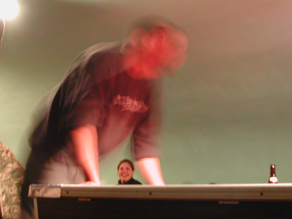 A person dancing with motion blur, leaning forward on a surface, while another person in the background smiles.