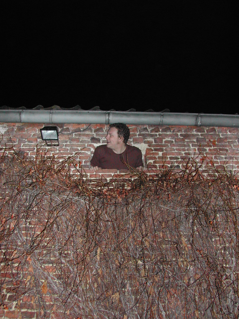 A man in a red shirt leans out of a small opening in a brick wall at night.