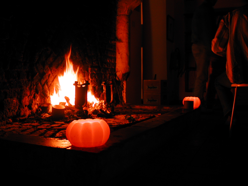 A fireplace with a burning fire, glowing pumpkin-shaped lights, and people sitting nearby in a dimly lit room.