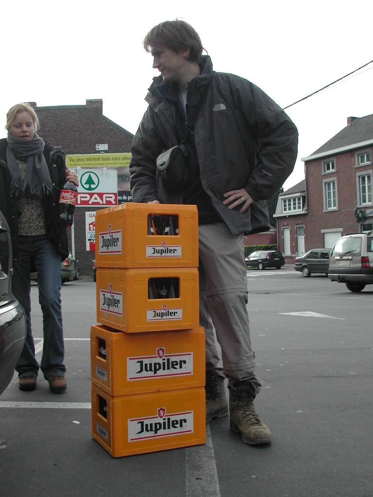 A man stands next to stacked beer crates, smiling, while a woman nearby holds a Coca-Cola bottle.