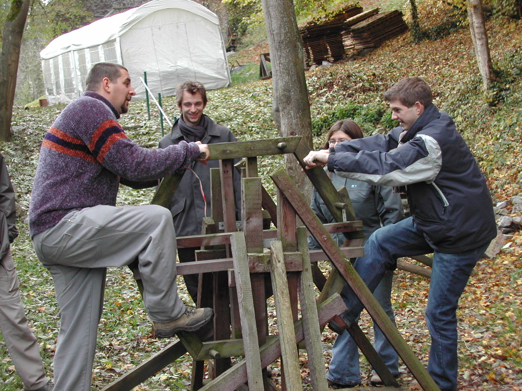 Four people outdoors interact with a wooden structure, two pushing or pulling while others observe and smile.
