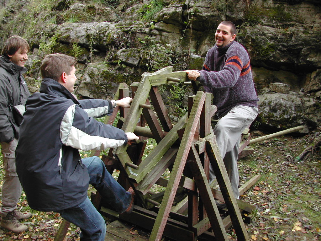 Two men play on a wooden seesaw-like structure while another person watches and smiles in an outdoor setting.