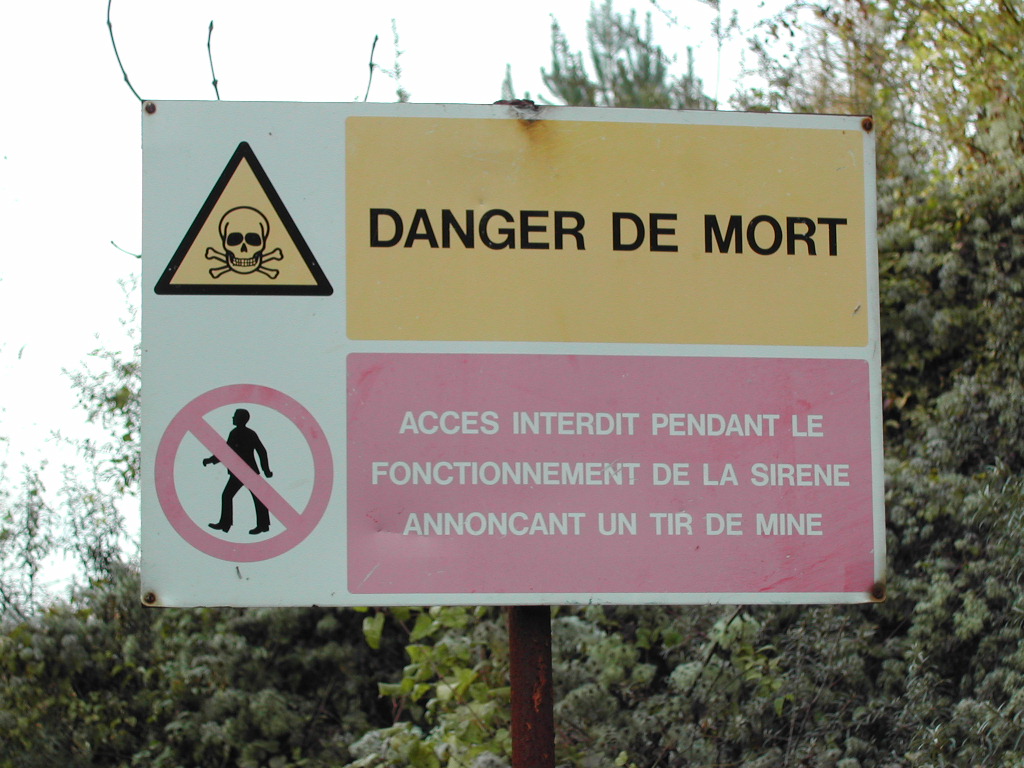 Warning sign with skull symbol reads "Danger de Mort", prohibiting access during siren operation for a mine blast.