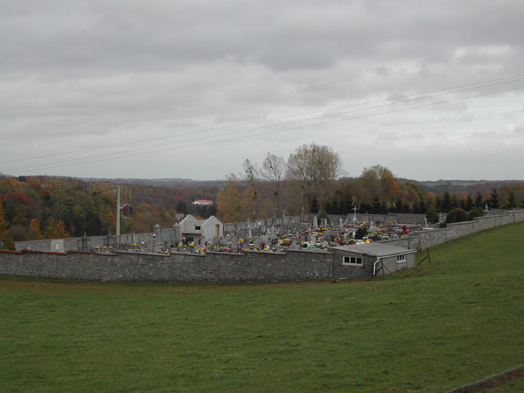 A cemetery with many graves decorated with flowers, viewed from a distance on a cloudy day.