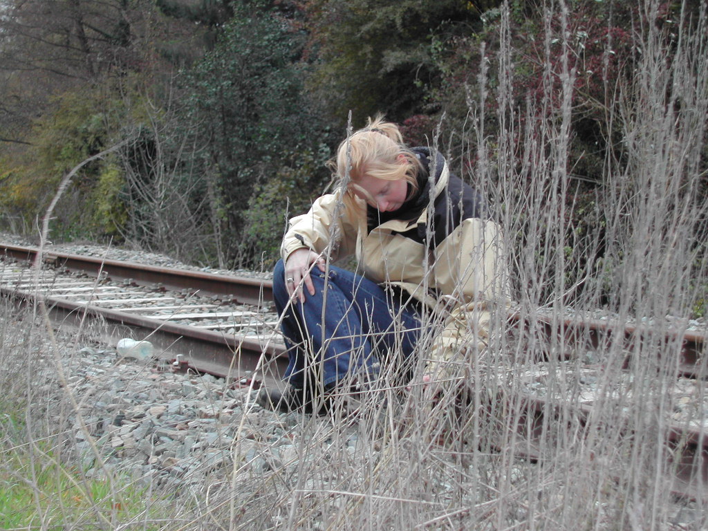 A person in a jacket crouches near railroad tracks, looking down and touching the ground.