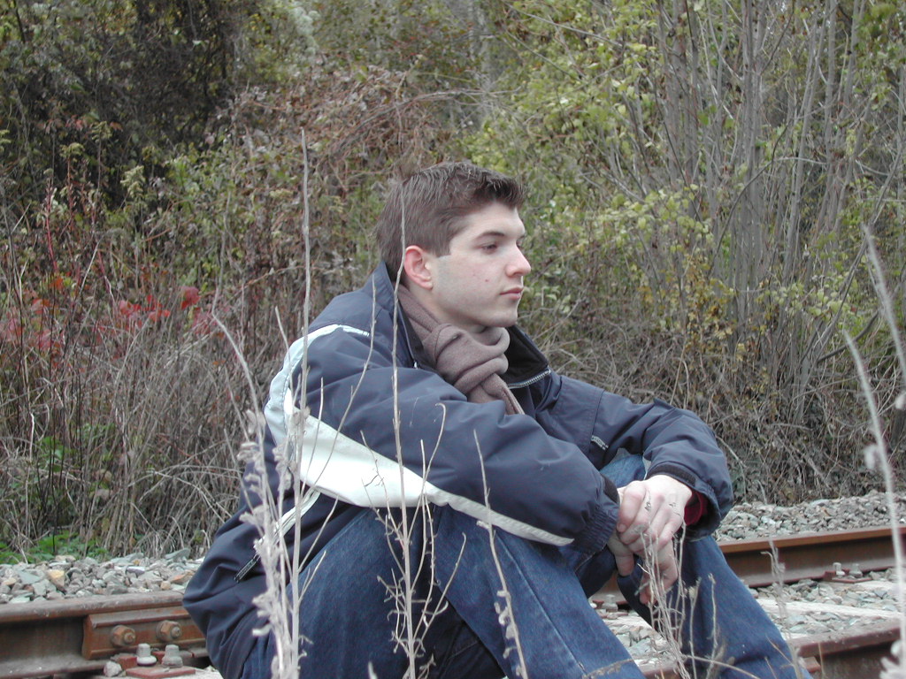 A person sits on the ground near train tracks, wearing a jacket and scarf, looking off into the distance.