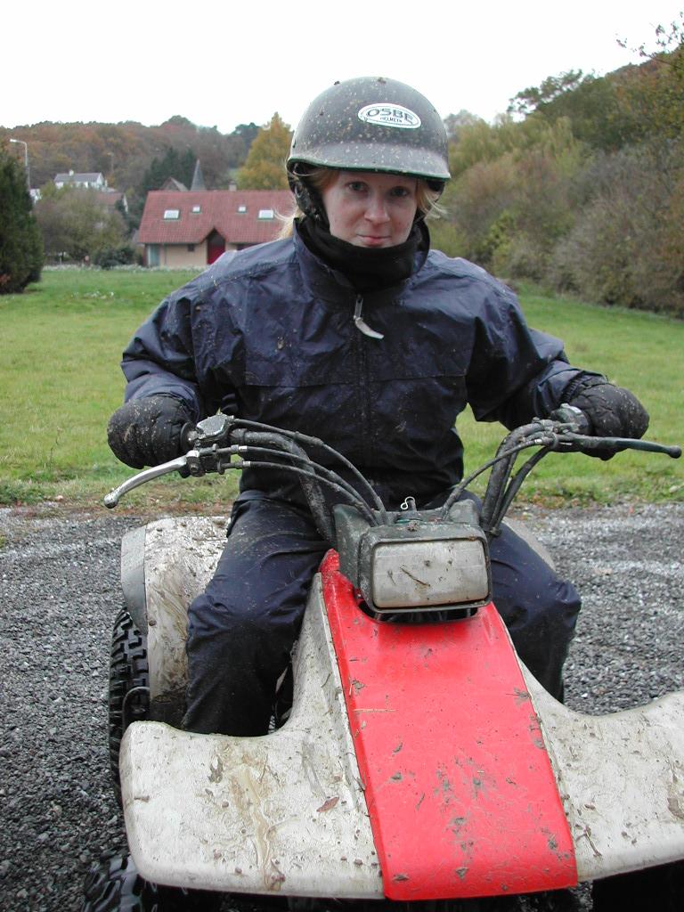 A person wearing a helmet and rain gear sits on a muddy all-terrain vehicle on a gravel path.