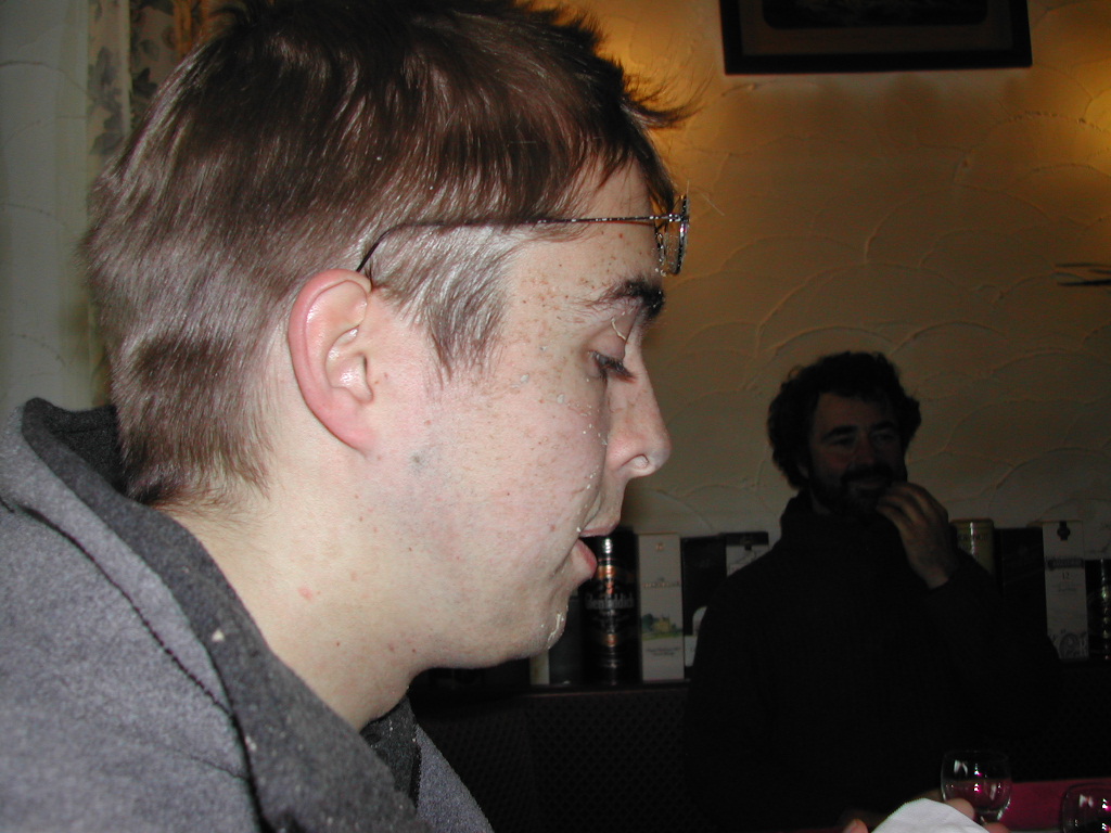 A man in profile with liquid on his face, wearing a dark hoodie, while another man smiles in the background.