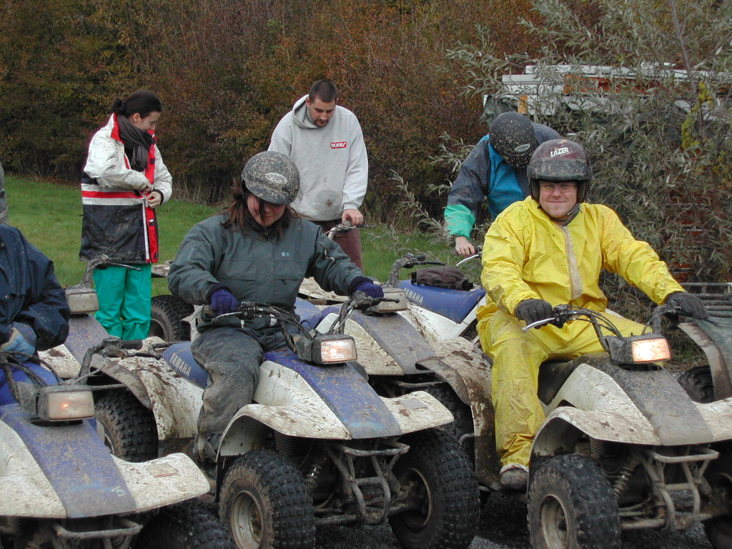 A group of people in helmets and muddy clothes ride all-terrain vehicles on a dirt path.