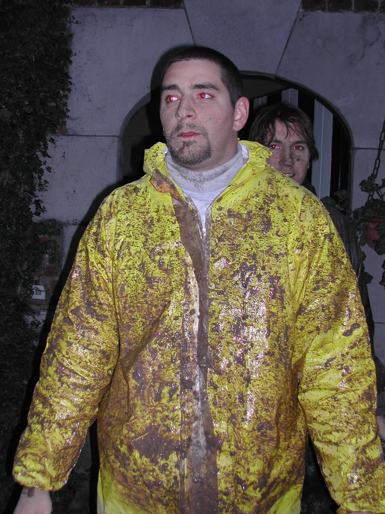 A man in a muddy yellow raincoat stands outside, with another person partially visible behind him.