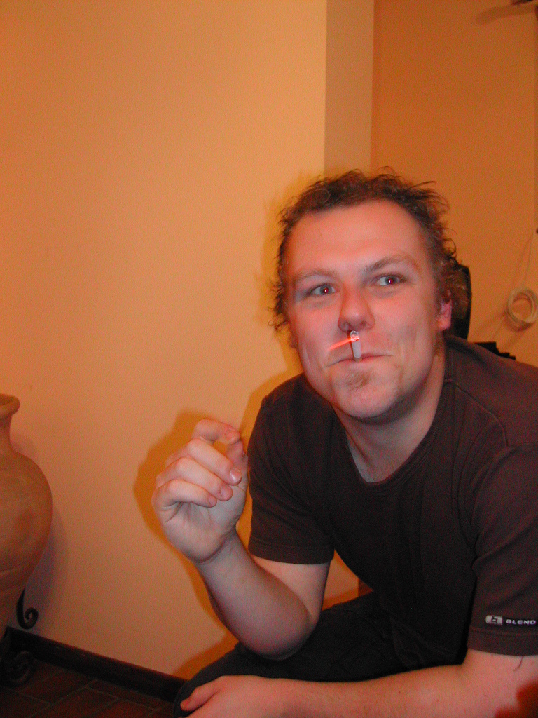 A man with a cigarette in his nose crouches indoors, looking at the camera with a playful expression.