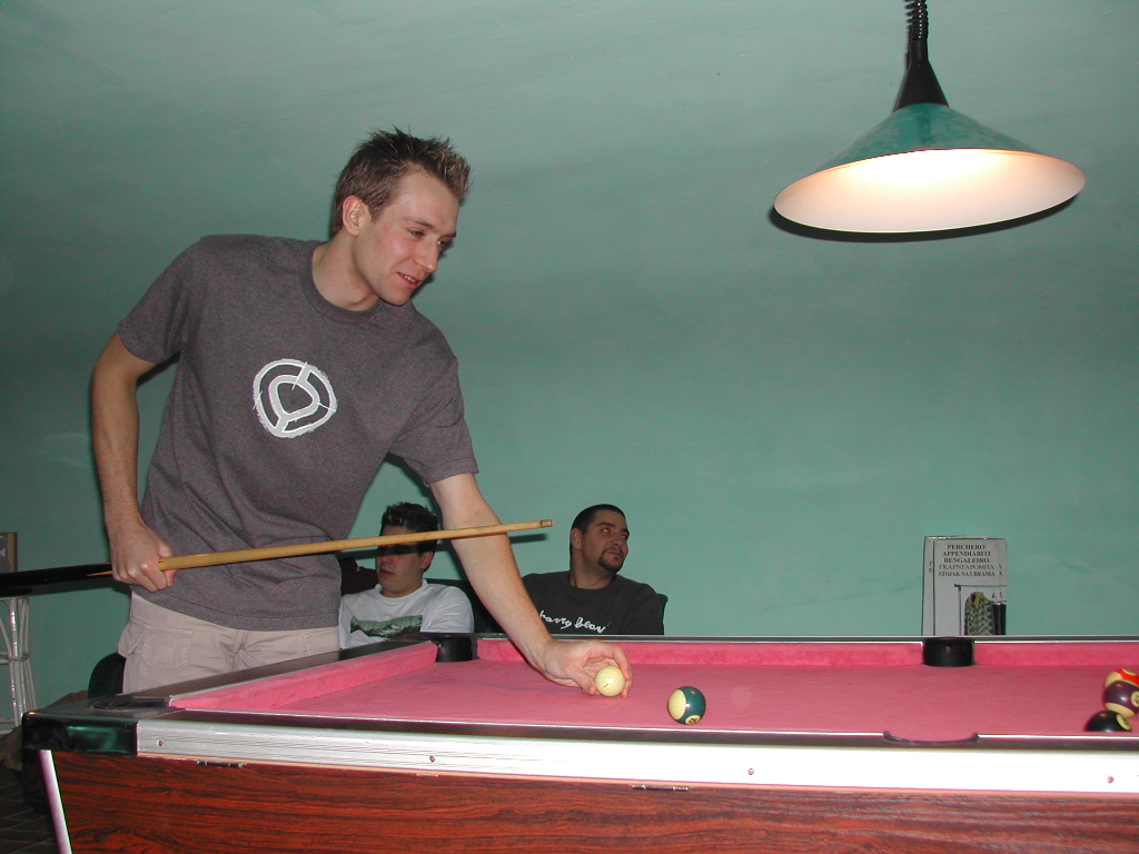 A man in a gray shirt prepares to take a shot at a pink pool table, holding a cue ball.