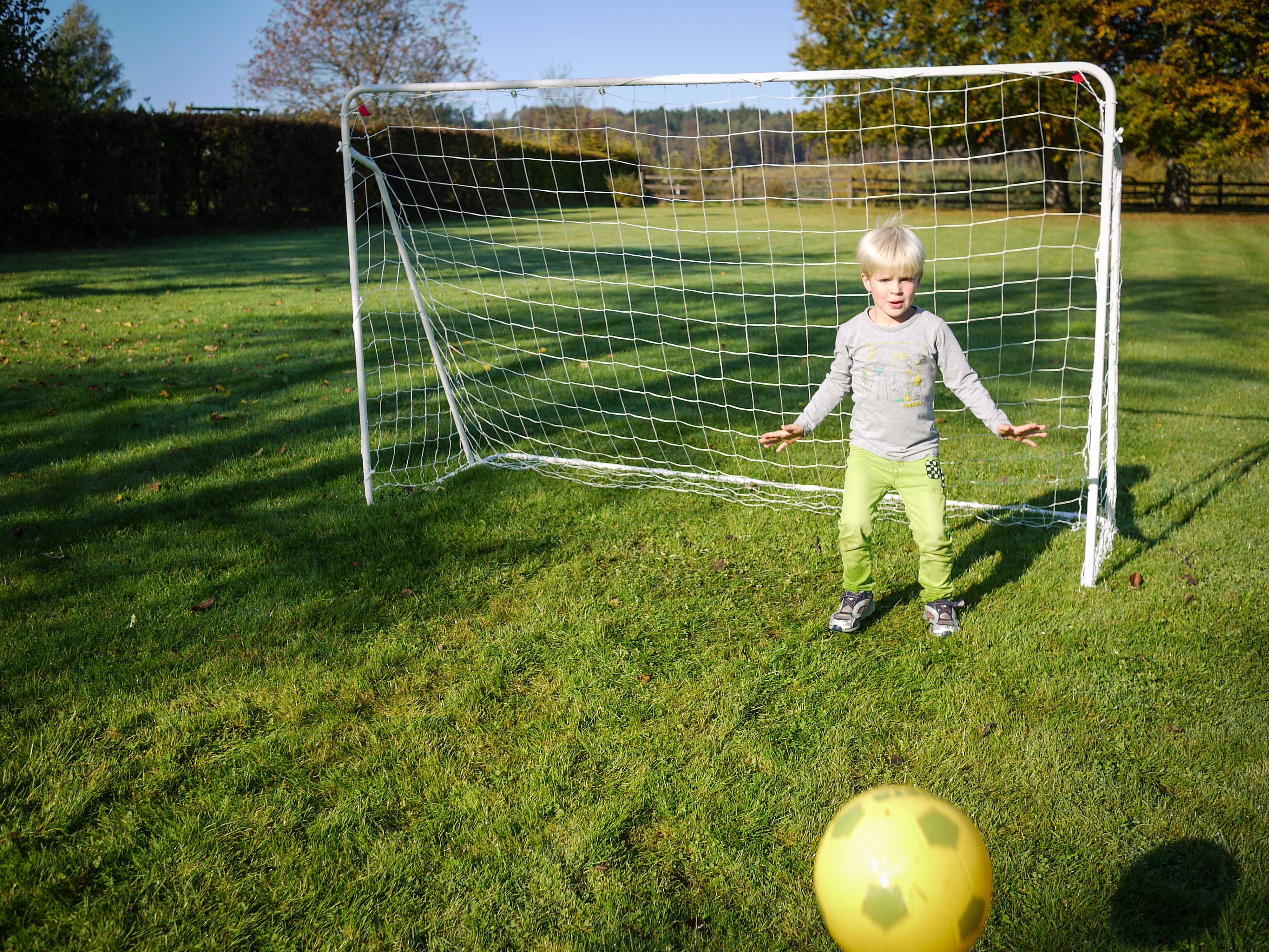 A child stands in front of a small soccer goal, preparing to block a yellow ball rolling toward them.