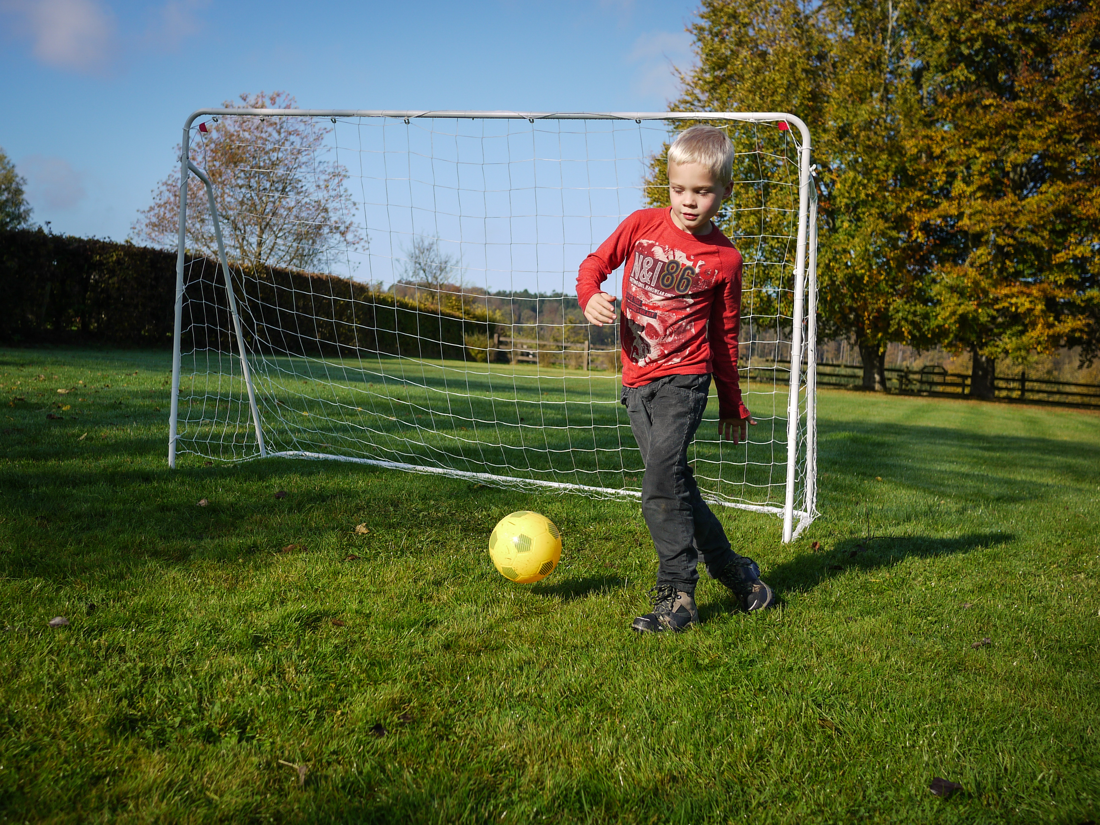 A child in a red shirt kicks a yellow soccer ball near a goal on a grassy field.