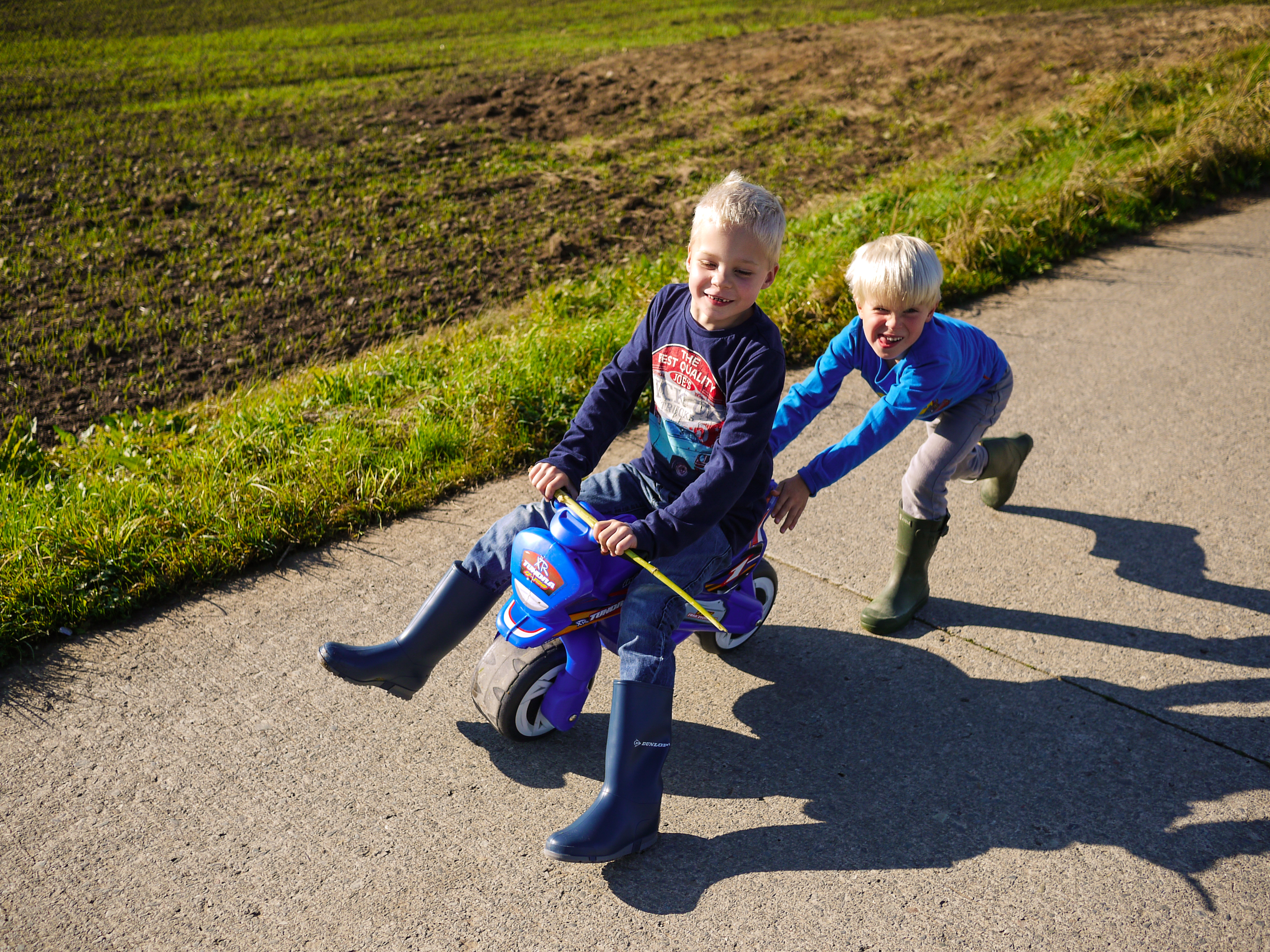 A child rides a small toy motorcycle while another child pushes from behind on a paved path.