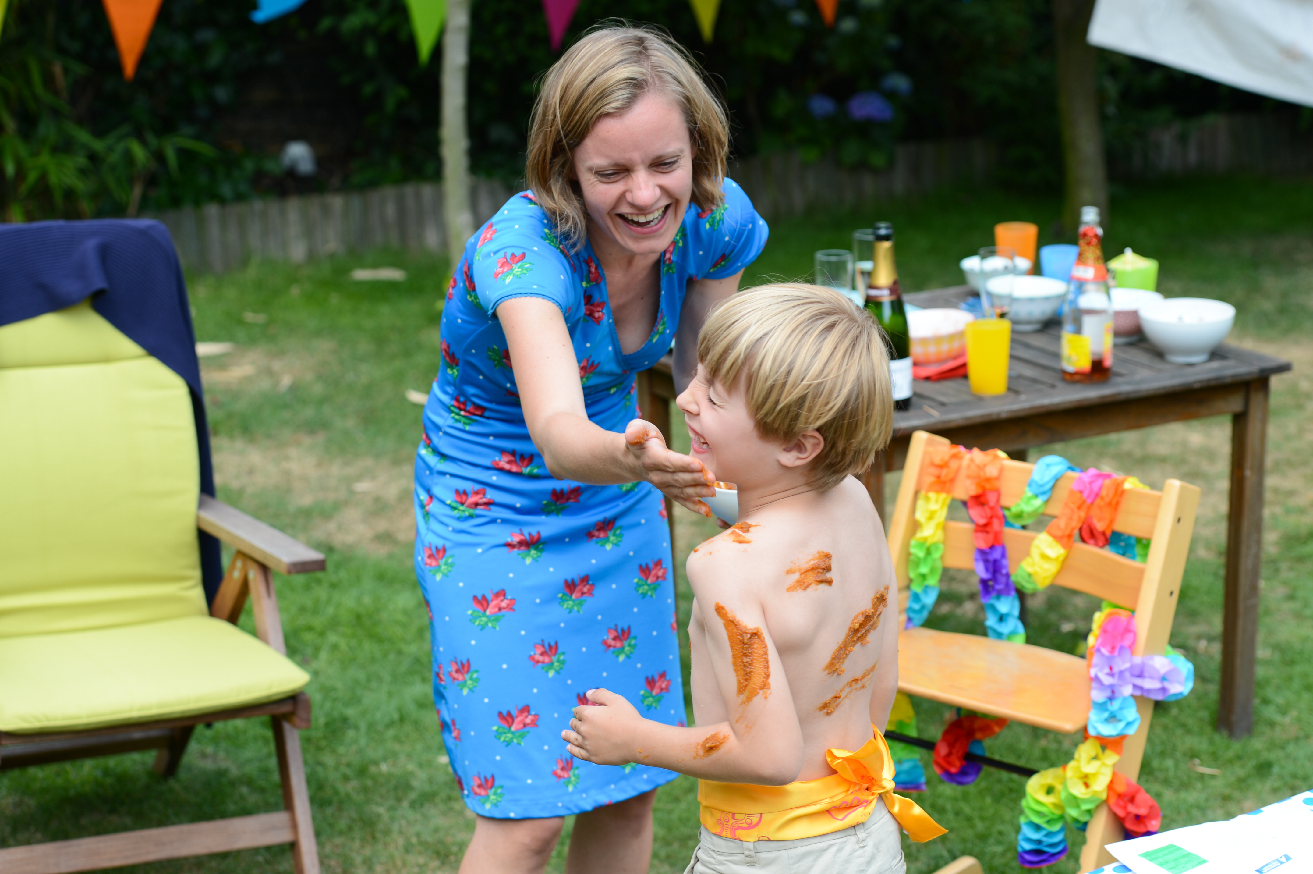 A smiling woman playfully spreads sauce on a laughing child's face and back at an outdoor celebration.