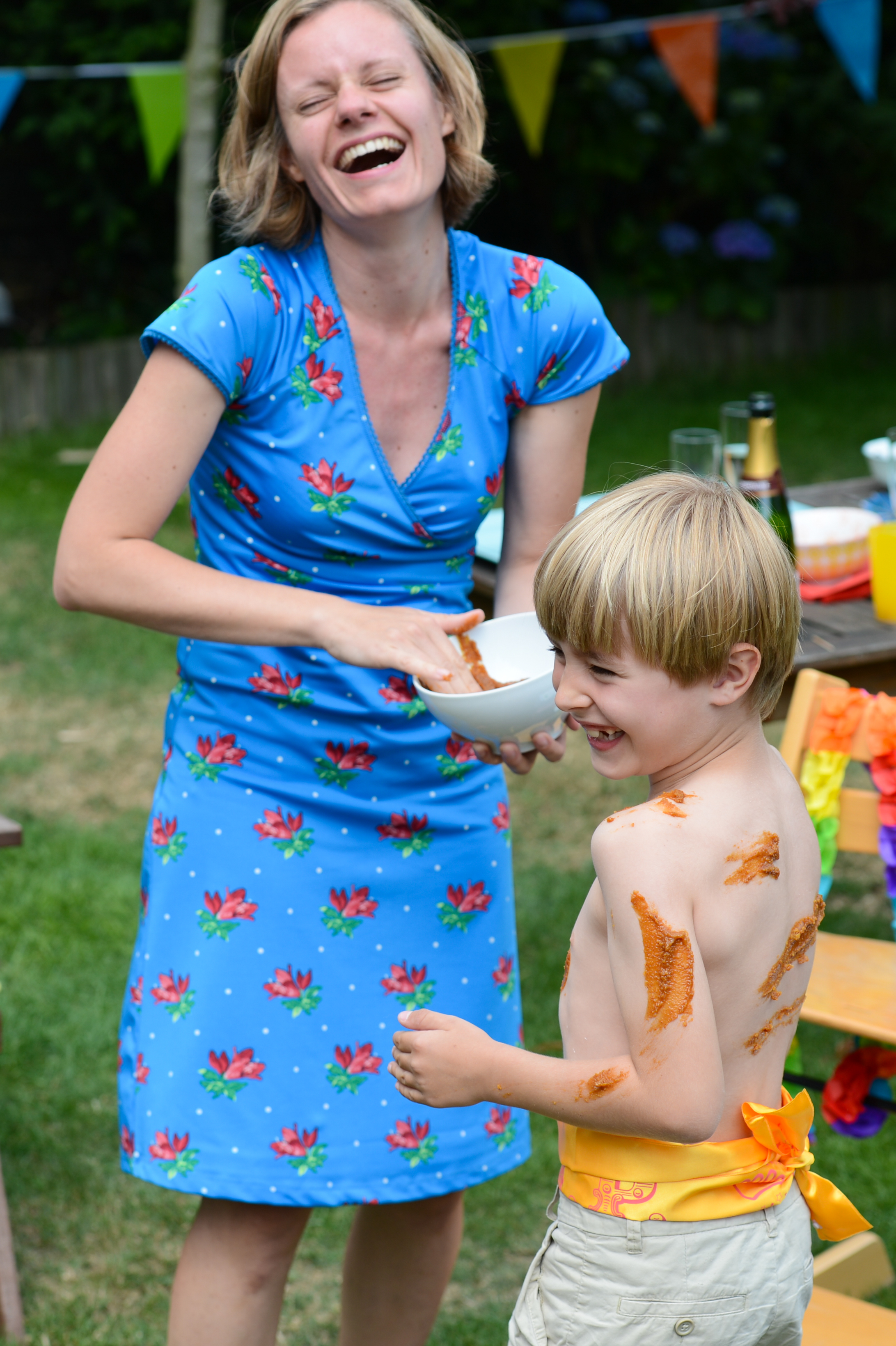 A woman laughs while applying sauce to a smiling child's bare back at an outdoor celebration.