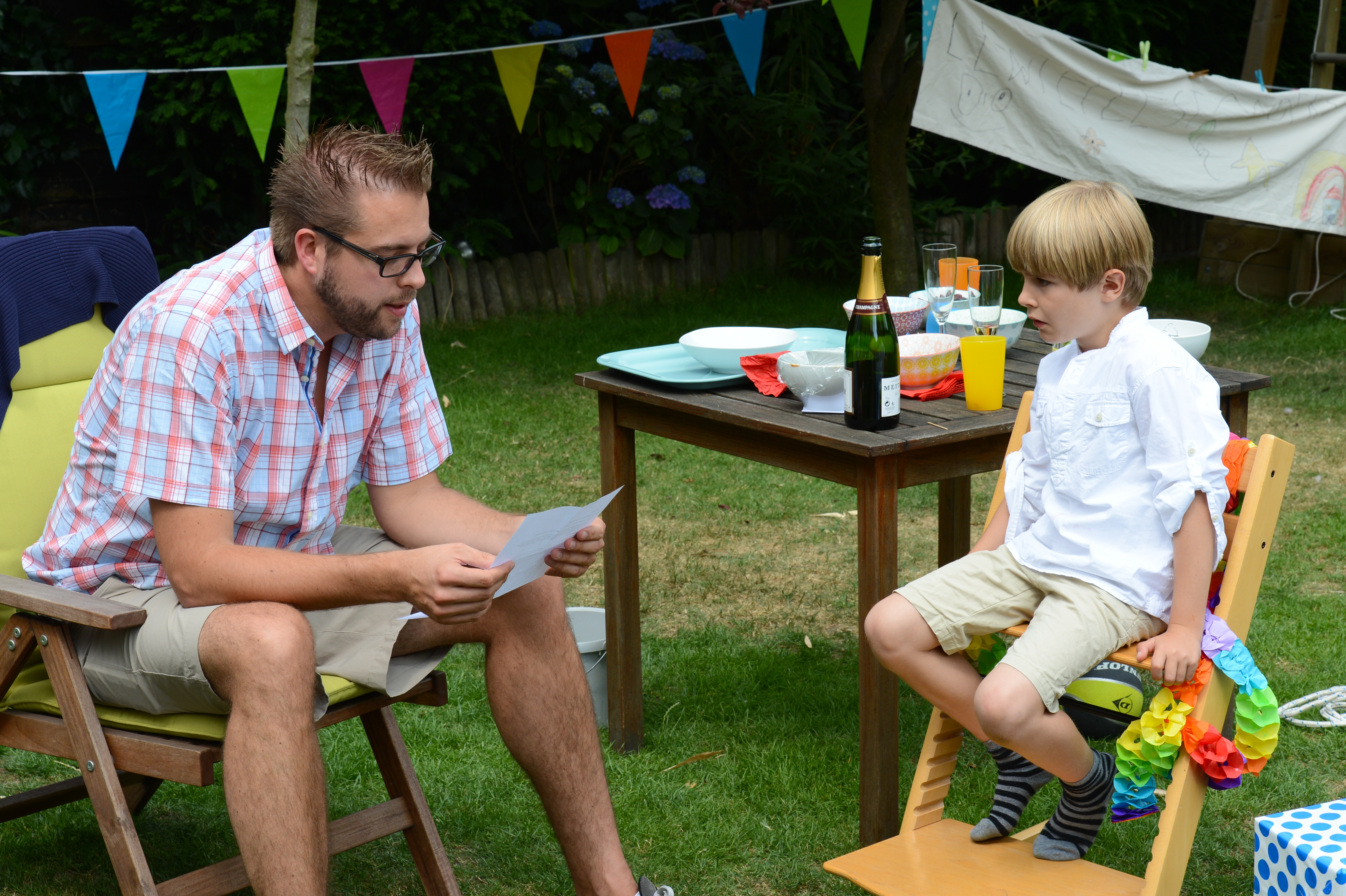 A man reads from a paper while a young boy in a decorated chair listens during an outdoor celebration.