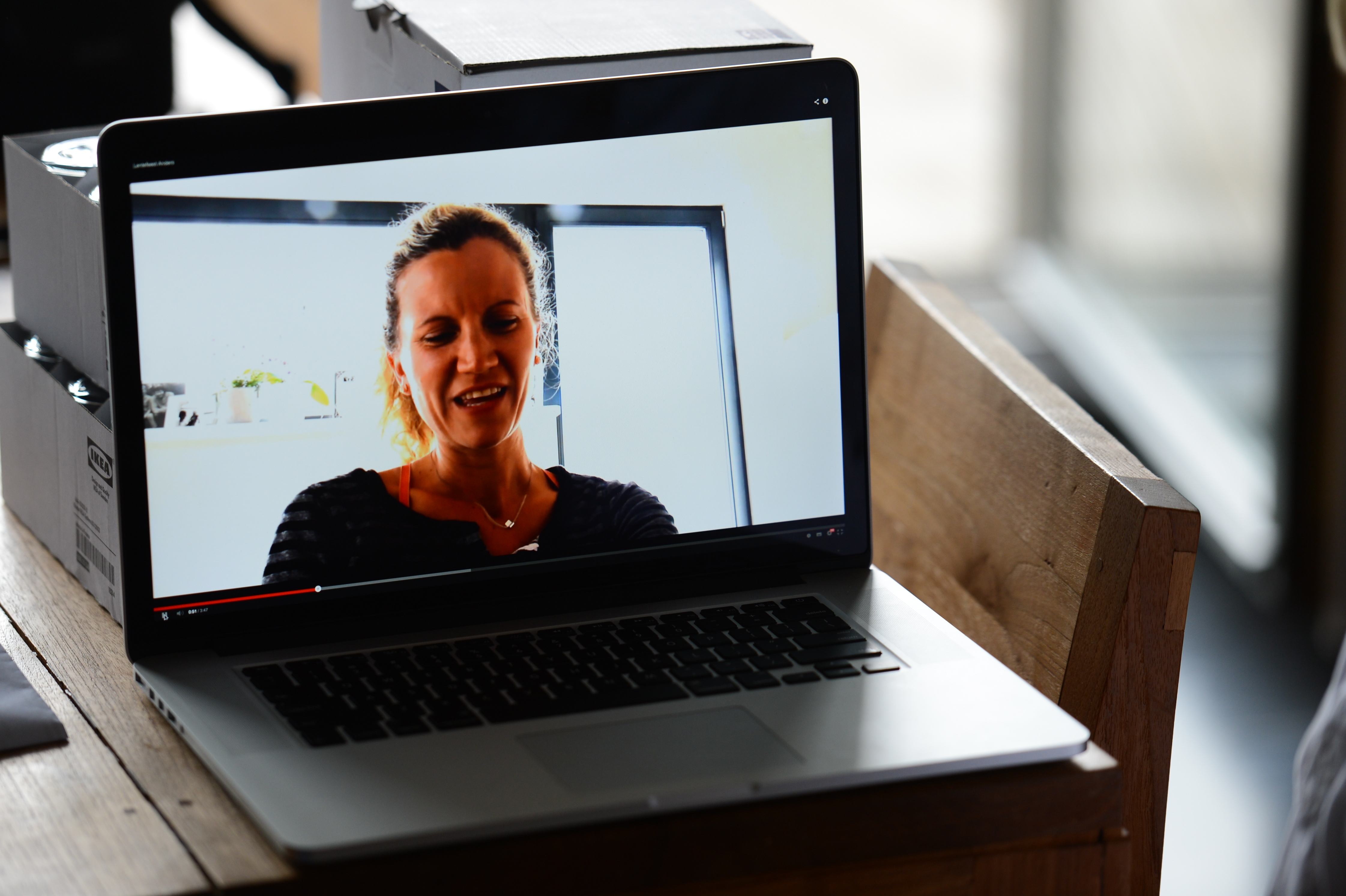 A laptop on a wooden table displays a video of a woman speaking in a well-lit room.