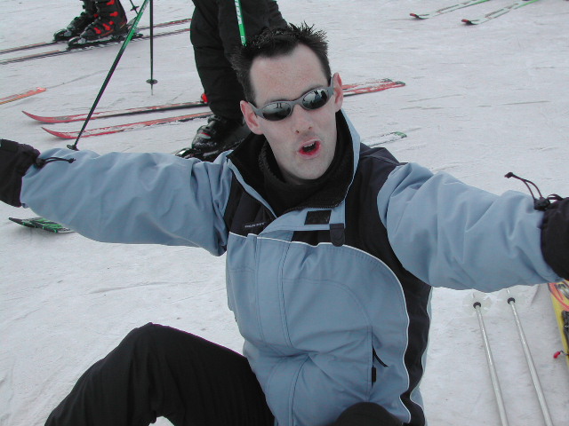 A person in a light blue ski jacket and sunglasses sits on the snow with arms outstretched, appearing excited.
