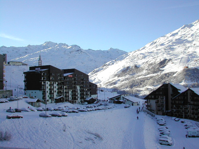 Snow-covered ski resort with buildings, parked cars, and people walking along a snowy path toward the slopes.