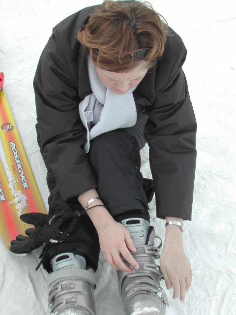 A person sitting on the snow fastens their ski boots, with a snowboard and gloves nearby.