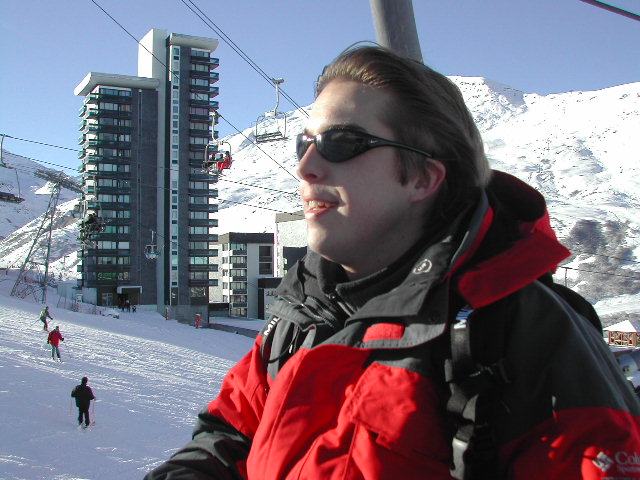 A person in a red and black jacket with sunglasses rides a ski lift in a snowy mountain resort.