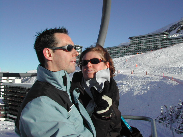 A man and woman wearing winter gear sit on a ski lift, surrounded by snowy slopes and buildings.